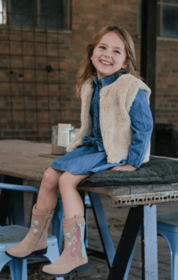 Hoe stijl je cowboylaarzen: meisje in spijkerjurk en teddybodywarmer met opvallende cowboylaarsjes in een rustige, stoere outfit.